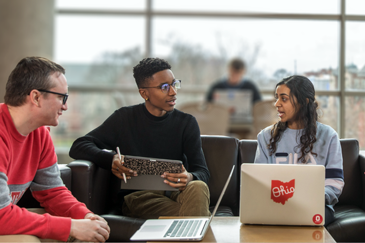 Three students with laptops discussing something while sitting in a library setting.
